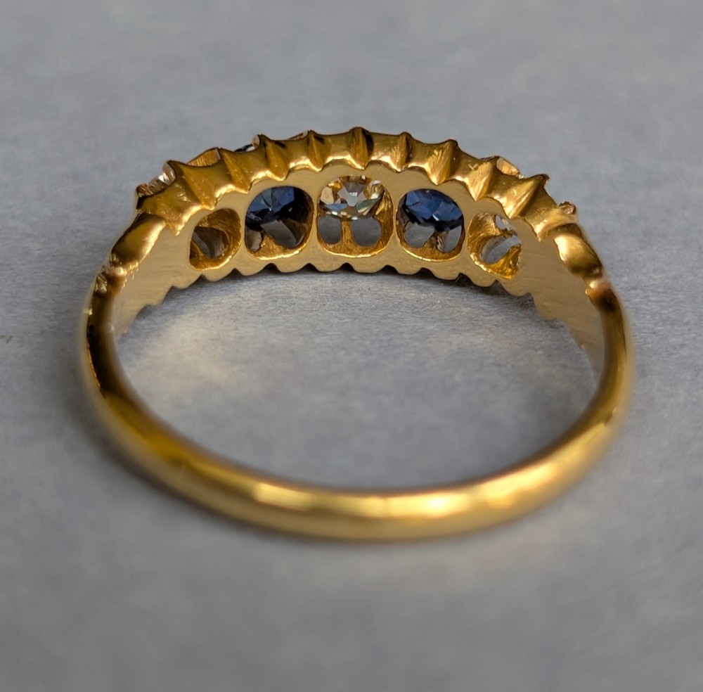 An Edwardian 18ct yellow gold five-stone ring featuring alternating blue sapphires and old cut diamonds set within a carved scroll head. View IX