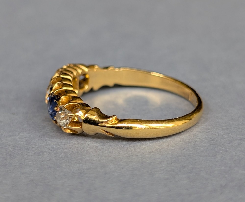 An Edwardian 18ct yellow gold five-stone ring featuring alternating blue sapphires and old cut diamonds set within a carved scroll head. View X