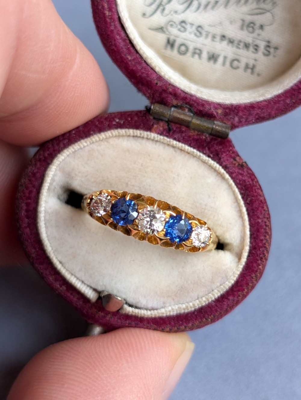 An Edwardian 18ct yellow gold five-stone ring featuring alternating blue sapphires and old cut diamonds set within a carved scroll head. View XI