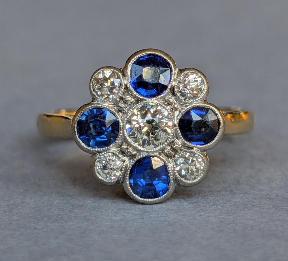 An Art Deco cluster ring featuring a central round diamond surrounded by alternating blue sapphires and smaller diamonds in a flower formation set on a gold band. View IV