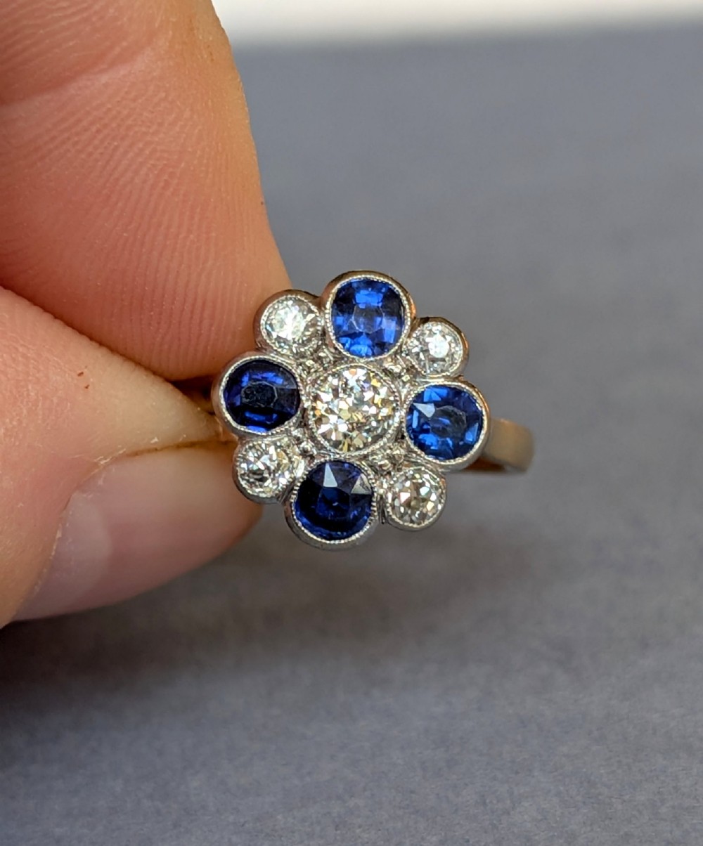 An Art Deco cluster ring featuring a central round diamond surrounded by alternating blue sapphires and smaller diamonds in a flower formation set on a gold band. View V