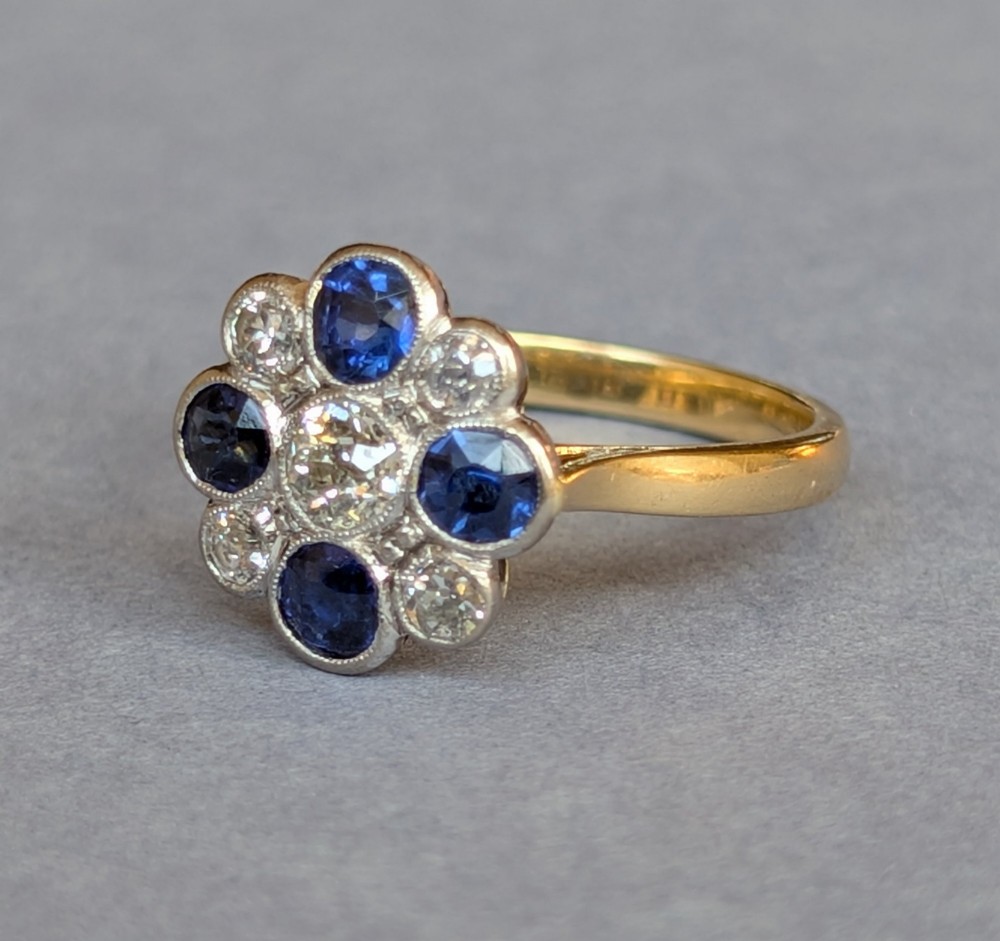 An Art Deco cluster ring featuring a central round diamond surrounded by alternating blue sapphires and smaller diamonds in a flower formation set on a gold band. View VI