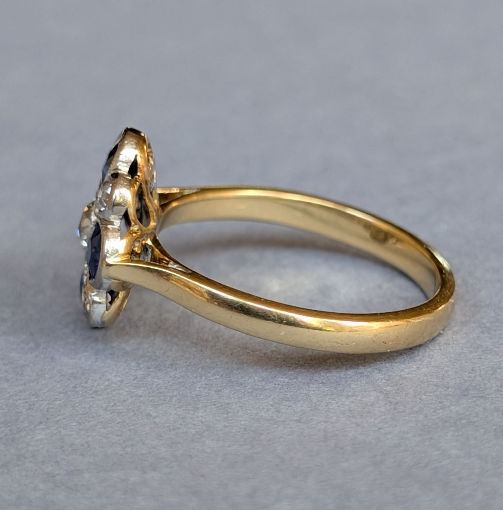 An Art Deco cluster ring featuring a central round diamond surrounded by alternating blue sapphires and smaller diamonds in a flower formation set on a gold band. View VII