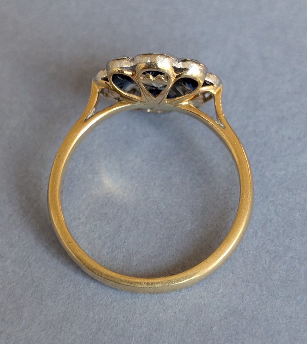 An Art Deco cluster ring featuring a central round diamond surrounded by alternating blue sapphires and smaller diamonds in a flower formation set on a gold band. View VIII