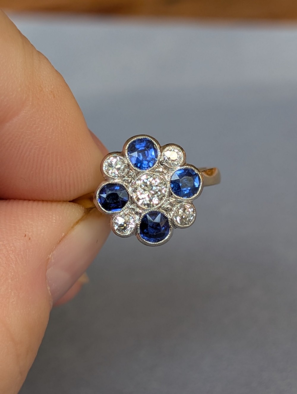 An Art Deco cluster ring featuring a central round diamond surrounded by alternating blue sapphires and smaller diamonds in a flower formation set on a gold band. View IX