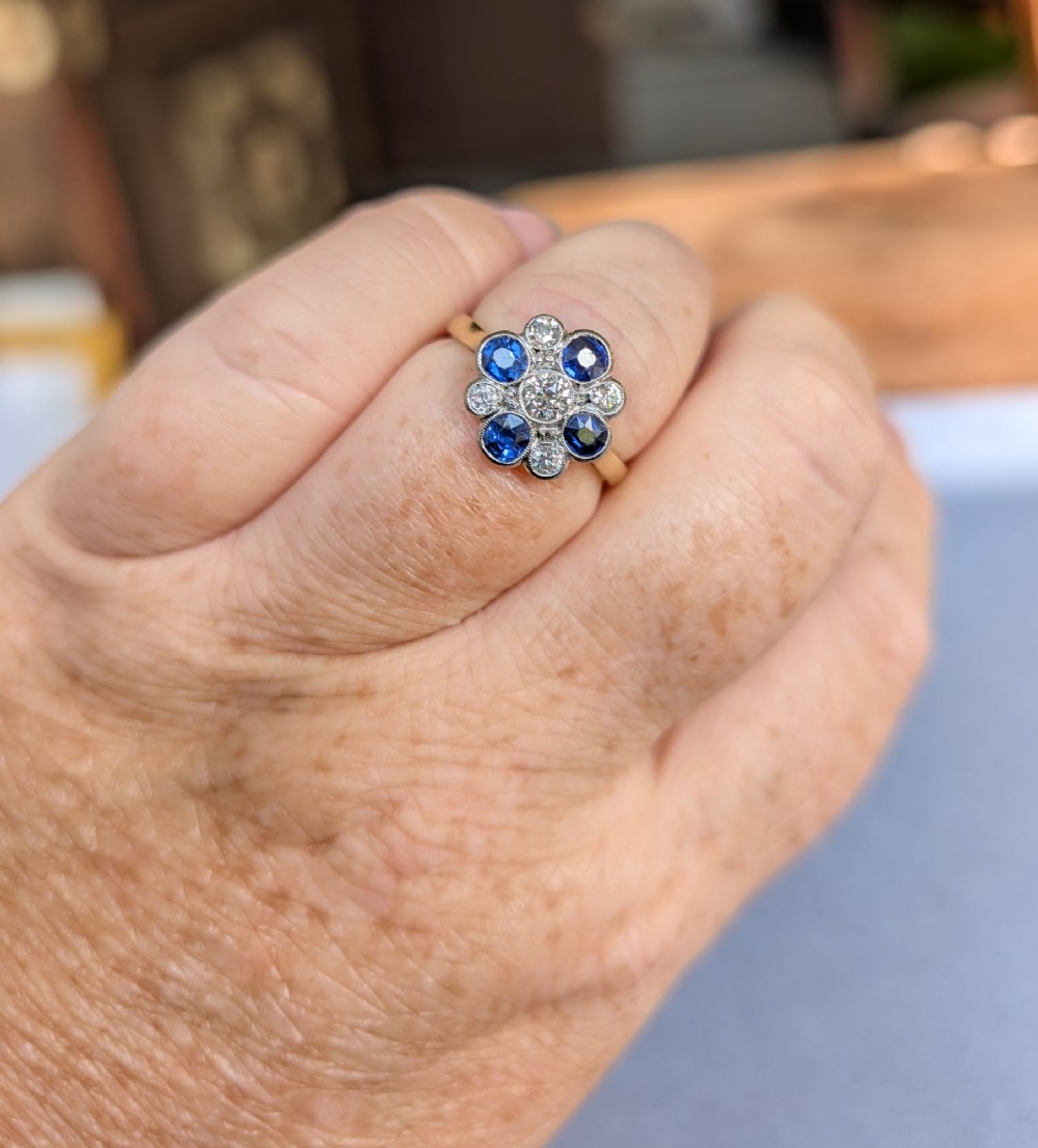 An Art Deco cluster ring featuring a central round diamond surrounded by alternating blue sapphires and smaller diamonds in a flower formation set on a gold band. View X