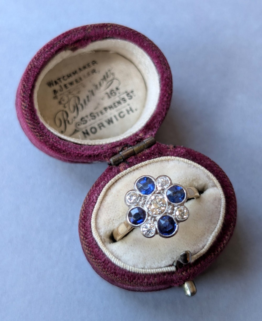 An Art Deco cluster ring featuring a central round diamond surrounded by alternating blue sapphires and smaller diamonds in a flower formation set on a gold band. View XII