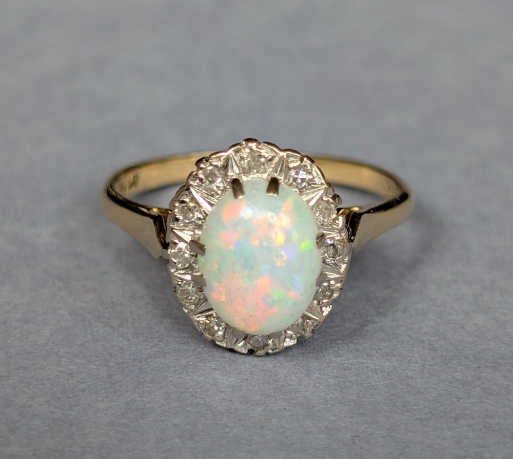 A mid-century 18ct gold ring featuring an oval cabochon opal framed by a halo cluster of small diamonds. View V