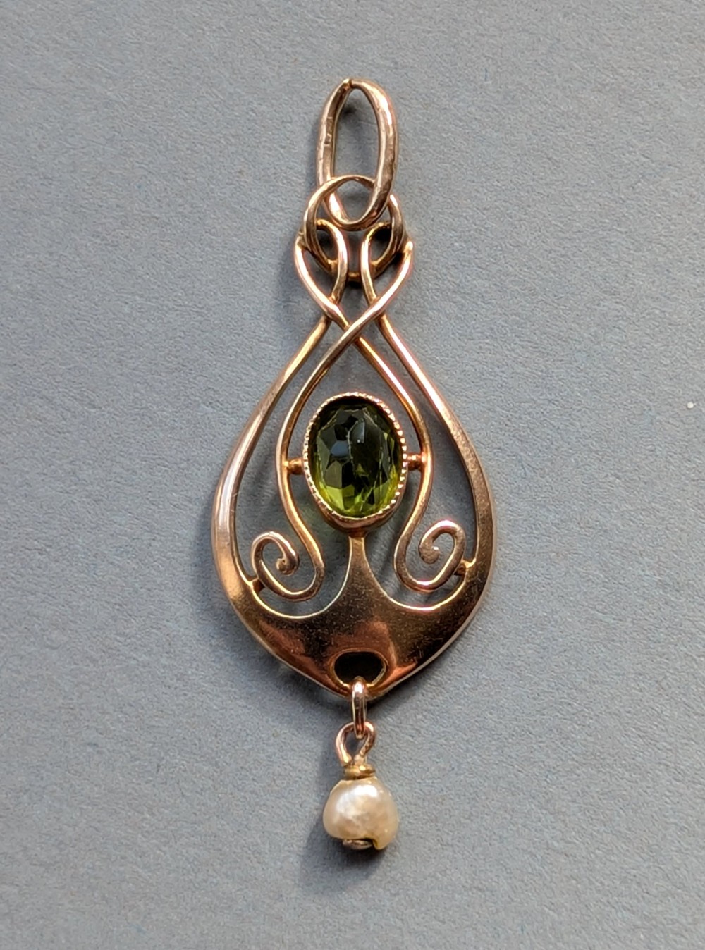 An Edwardian 9ct gold openwork Art Nouveau pendant featuring an oval bezel-set green peridot and a suspended pearl drop.