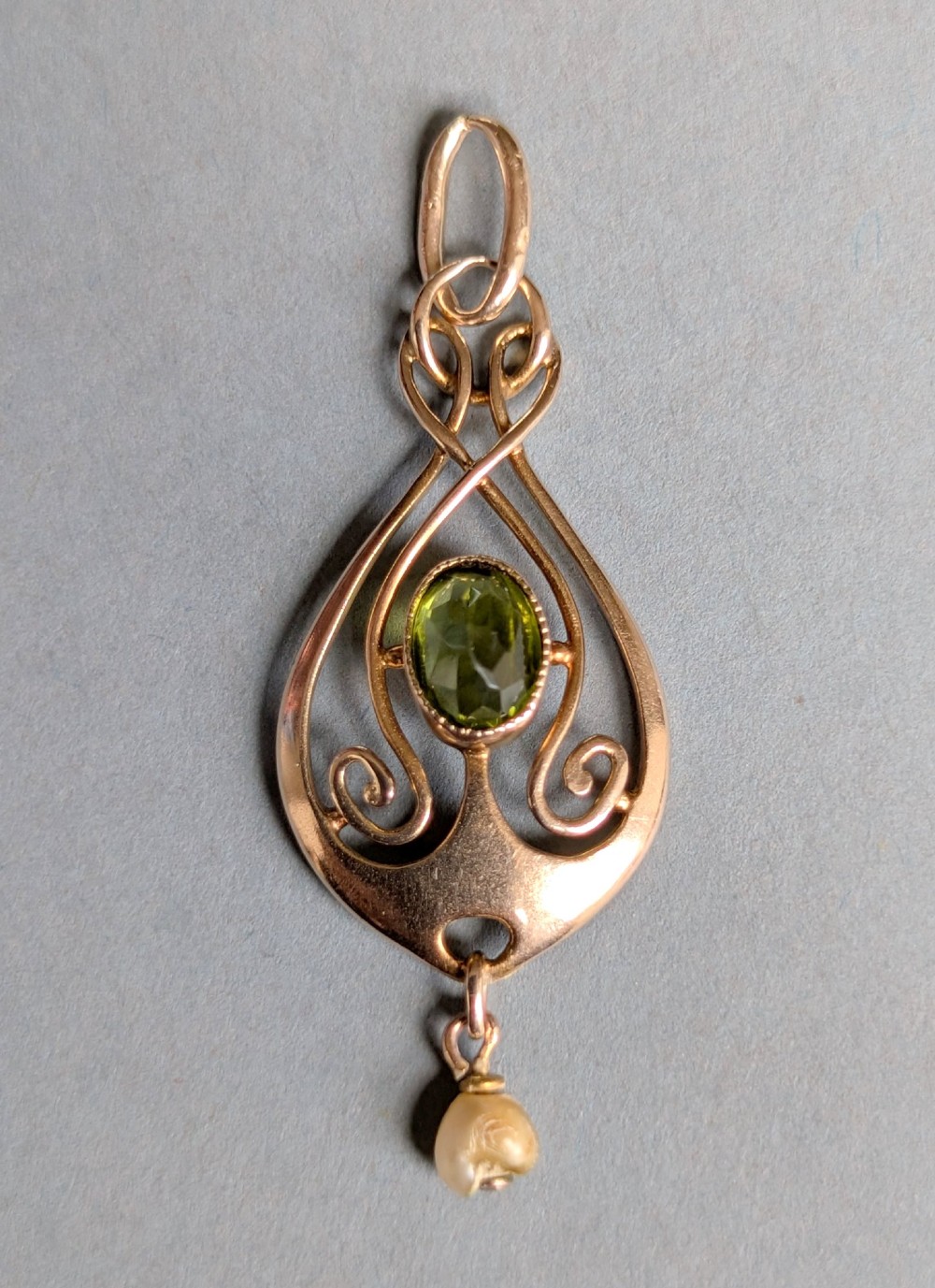 An Edwardian 9ct gold openwork Art Nouveau pendant featuring an oval bezel-set green peridot and a suspended pearl drop. View V
