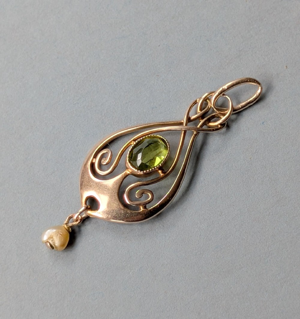 An Edwardian 9ct gold openwork Art Nouveau pendant featuring an oval bezel-set green peridot and a suspended pearl drop. View X
