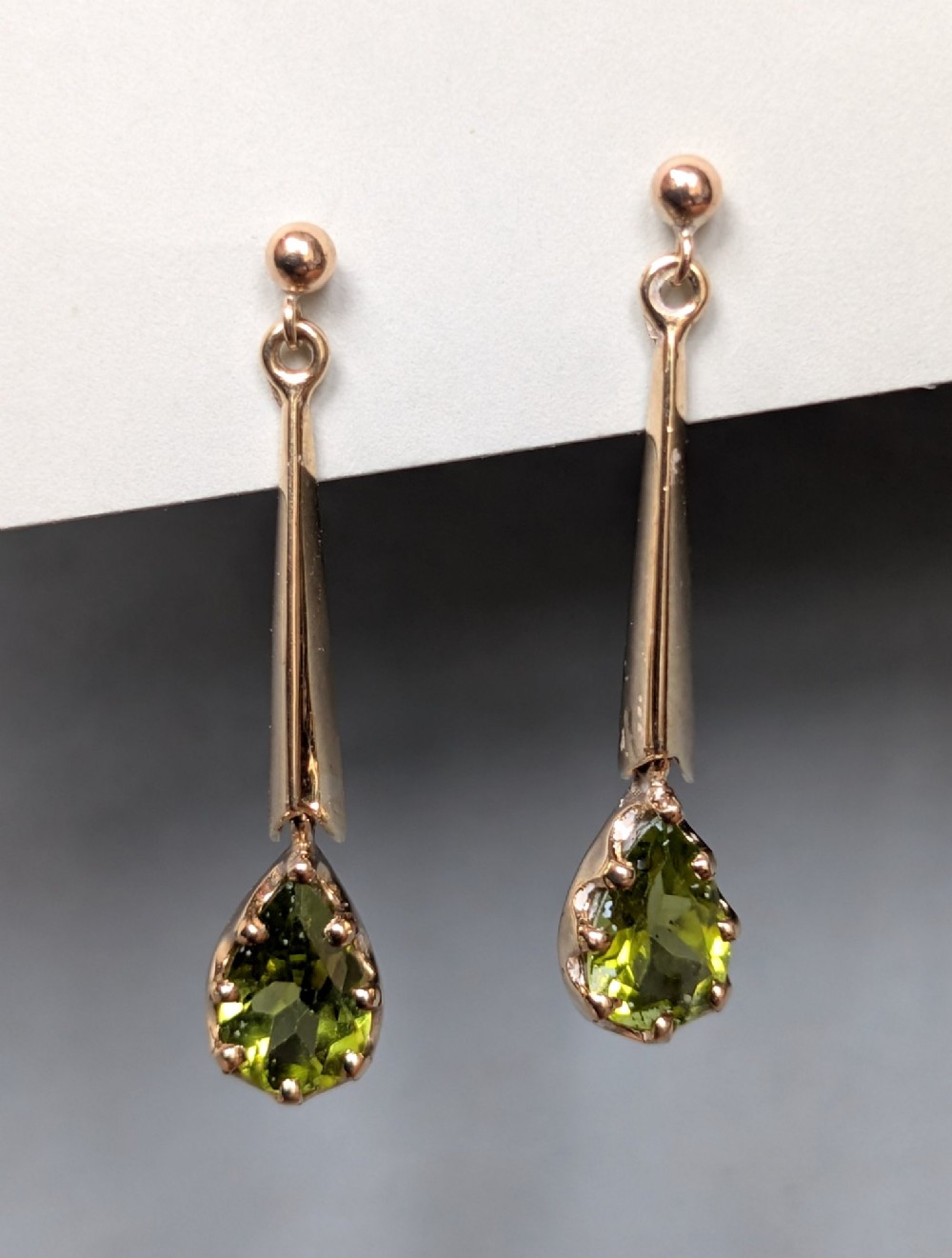 A pair of antique 9ct gold drop earrings featuring tapered bars and pear-cut green peridot gemstones suspended from ball posts.