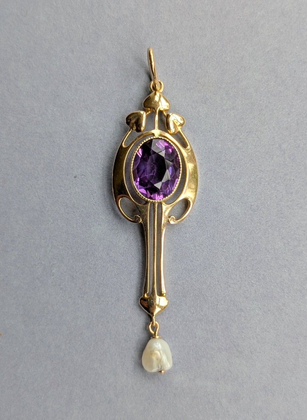An Art Nouveau 15ct gold drop pendant featuring a central oval faceted purple amethyst framed by stylised leaf motifs with a suspended pearl at the base.