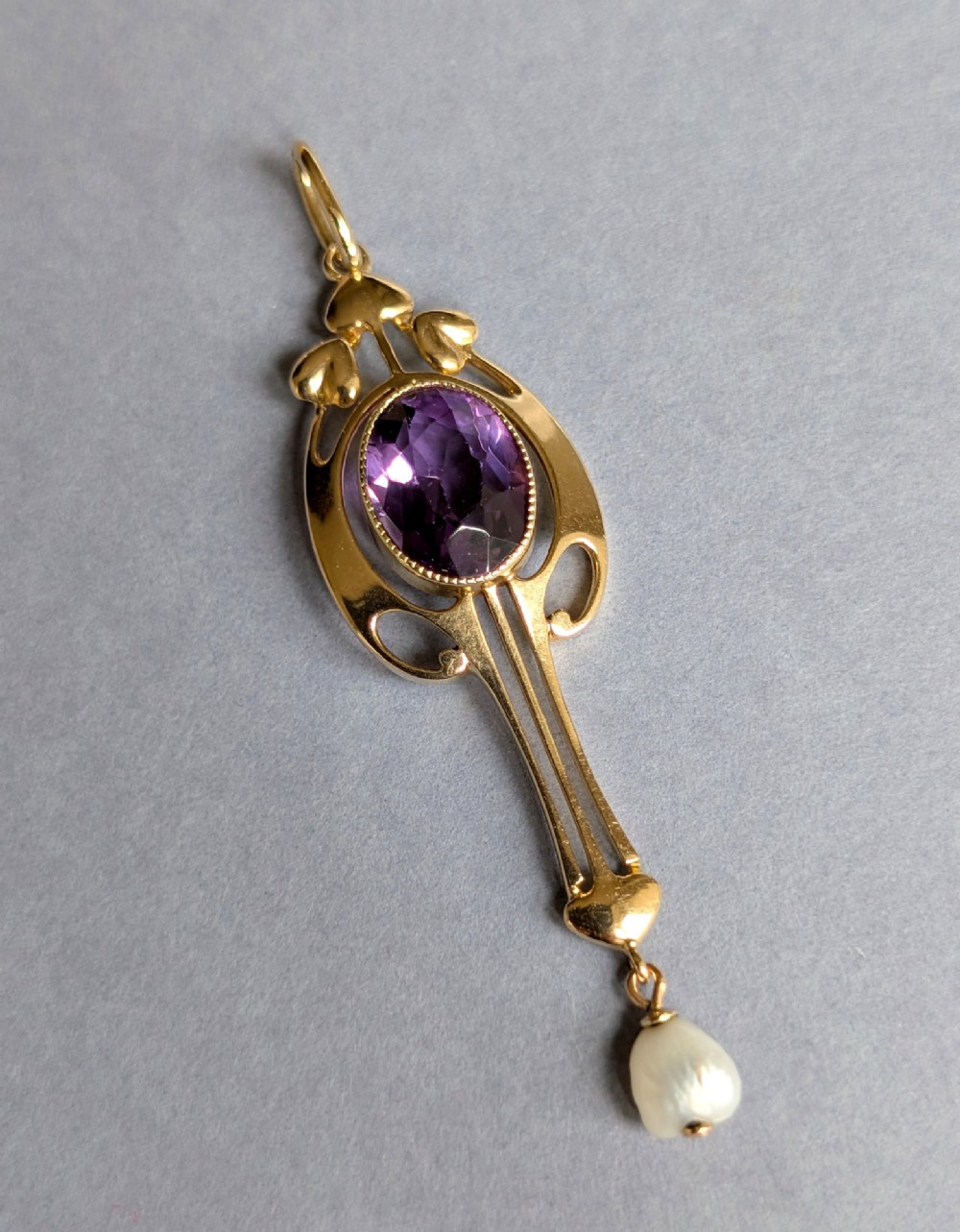 An Art Nouveau 15ct gold drop pendant featuring a central oval faceted purple amethyst framed by stylised leaf motifs with a suspended pearl at the base. View V