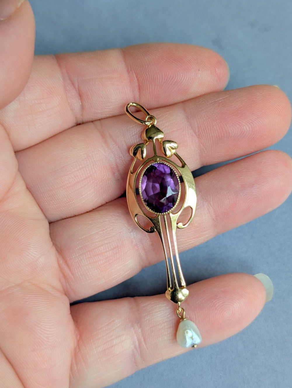 An Art Nouveau 15ct gold drop pendant featuring a central oval faceted purple amethyst framed by stylised leaf motifs with a suspended pearl at the base. View VII