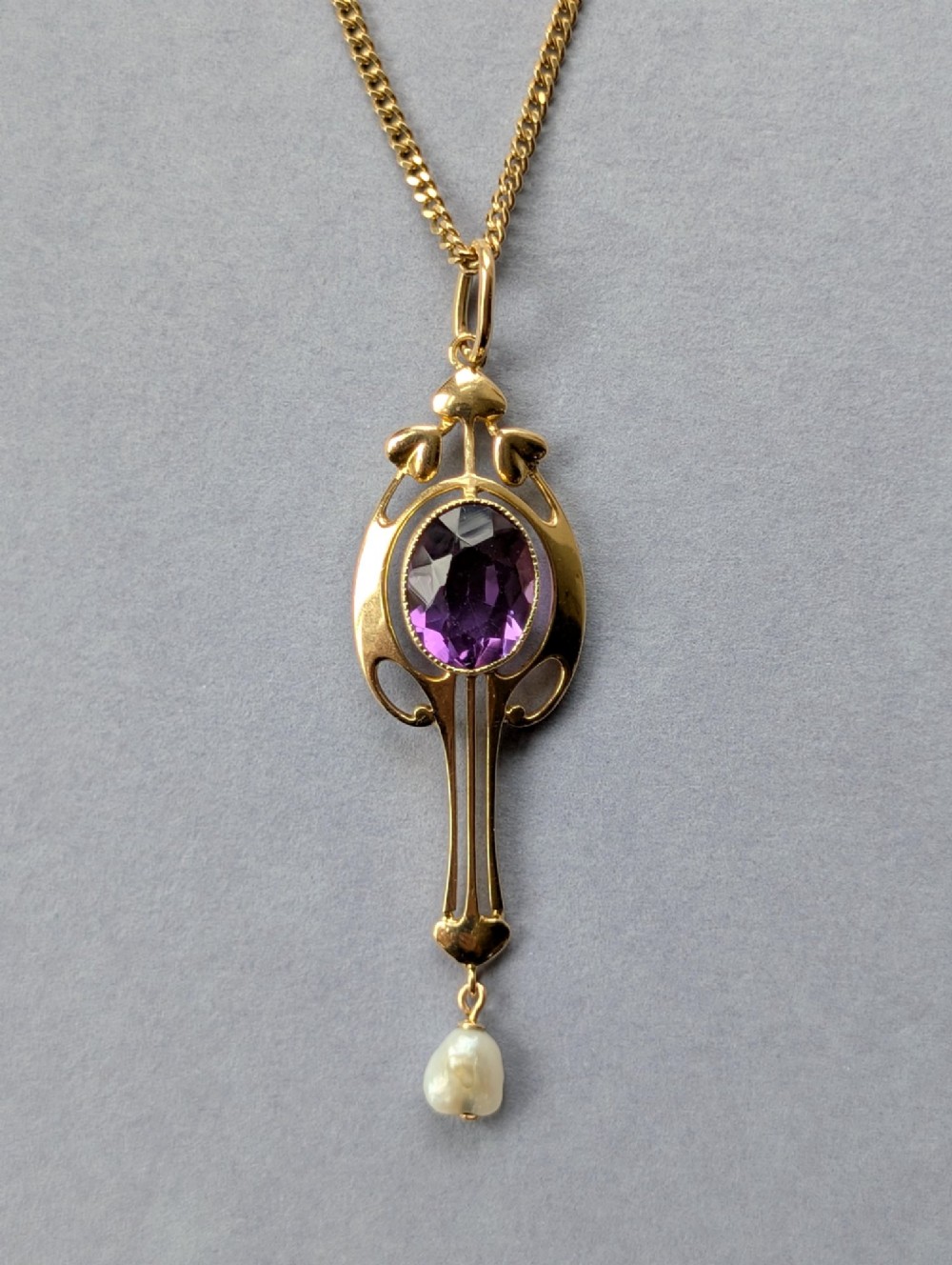 An Art Nouveau 15ct gold drop pendant featuring a central oval faceted purple amethyst framed by stylised leaf motifs with a suspended pearl at the base. View XIII