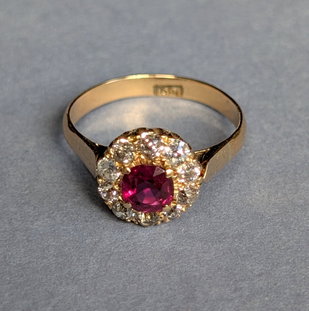 An antique 18ct gold cluster ring featuring a central faceted red ruby surrounded by ten round diamonds. View X