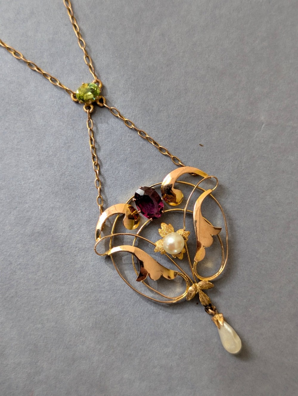 An Edwardian openwork gold lavalier pendant set with an oval purple garnet, a seed pearl center, a pearl drop, and a small green peridot, suspended from a gold chain. View II