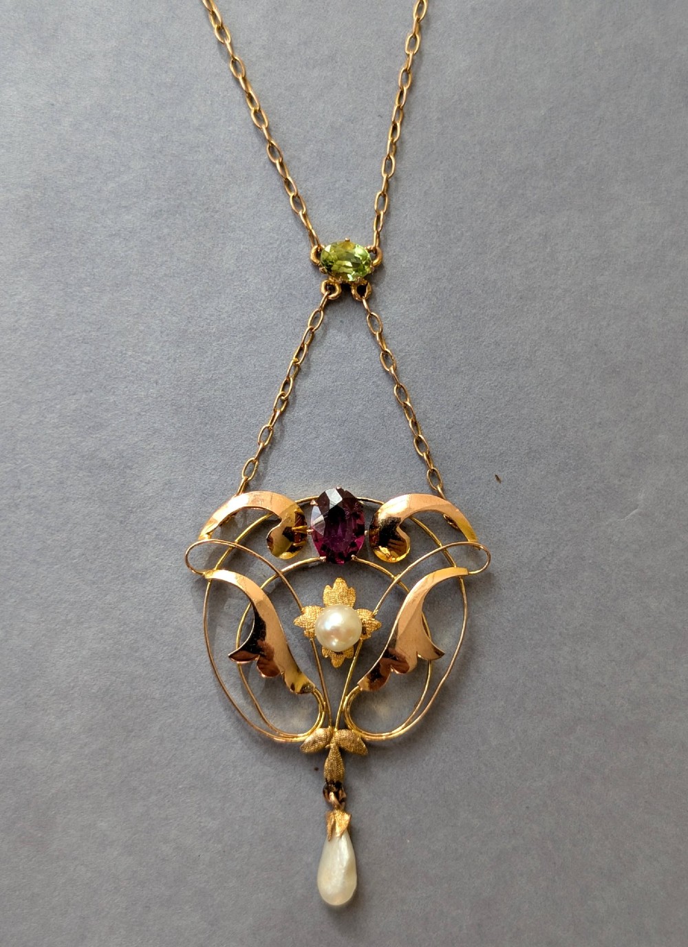 An Edwardian openwork gold lavalier pendant set with an oval purple garnet, a seed pearl center, a pearl drop, and a small green peridot, suspended from a gold chain.