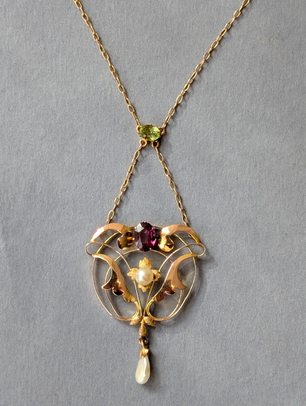 An Edwardian openwork gold lavalier pendant set with an oval purple garnet, a seed pearl center, a pearl drop, and a small green peridot, suspended from a gold chain. View IV