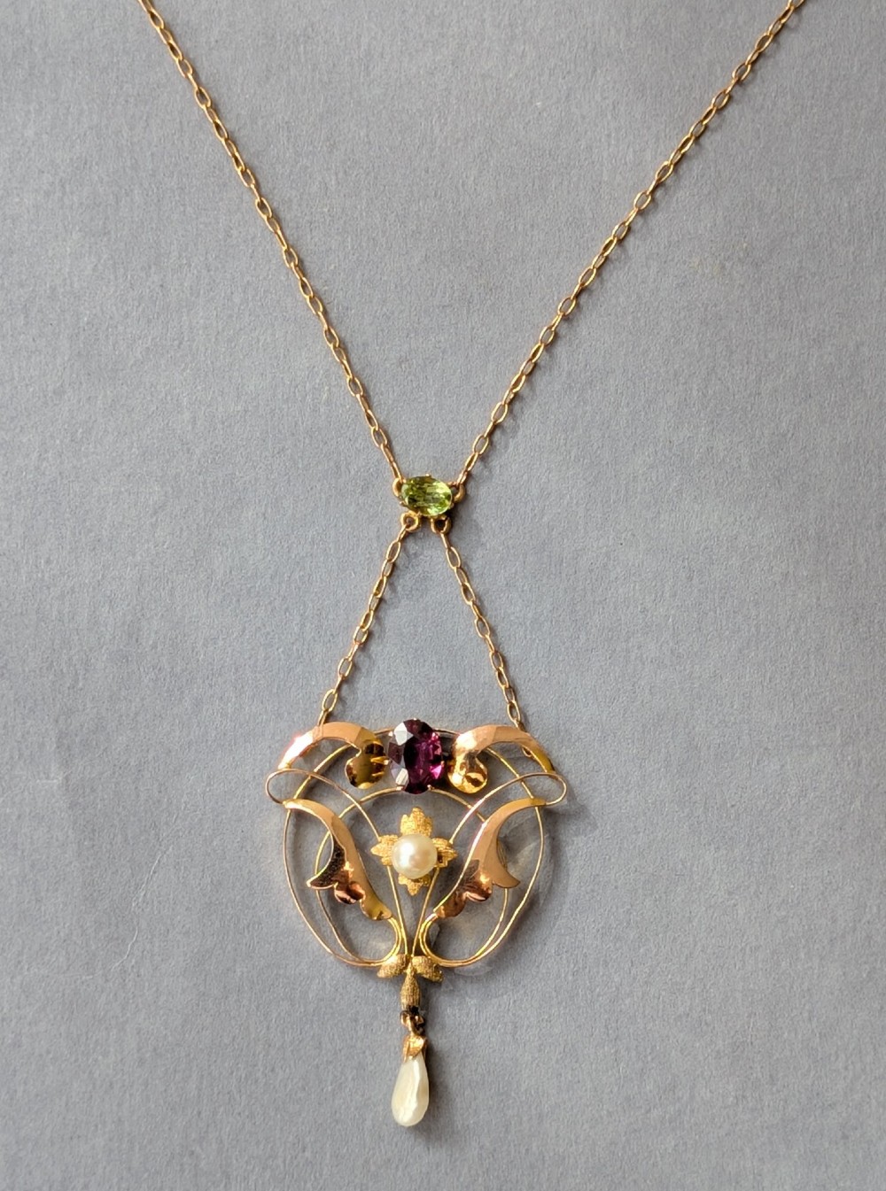 An Edwardian openwork gold lavalier pendant set with an oval purple garnet, a seed pearl center, a pearl drop, and a small green peridot, suspended from a gold chain. View V