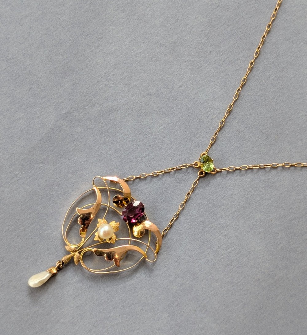 An Edwardian openwork gold lavalier pendant set with an oval purple garnet, a seed pearl center, a pearl drop, and a small green peridot, suspended from a gold chain. View VI