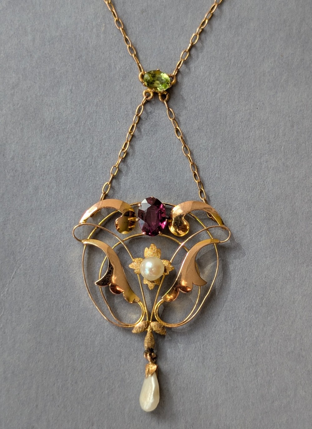 An Edwardian openwork gold lavalier pendant set with an oval purple garnet, a seed pearl center, a pearl drop, and a small green peridot, suspended from a gold chain. View VII