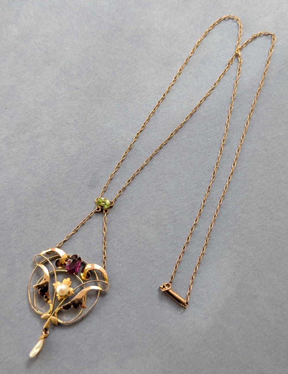 An Edwardian openwork gold lavalier pendant set with an oval purple garnet, a seed pearl center, a pearl drop, and a small green peridot, suspended from a gold chain. View X