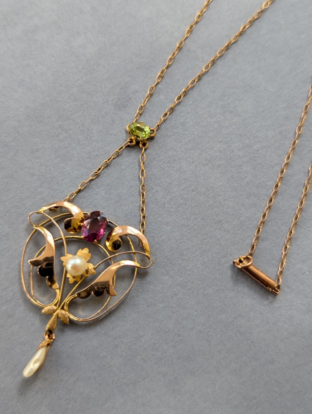 An Edwardian openwork gold lavalier pendant set with an oval purple garnet, a seed pearl center, a pearl drop, and a small green peridot, suspended from a gold chain. View XI