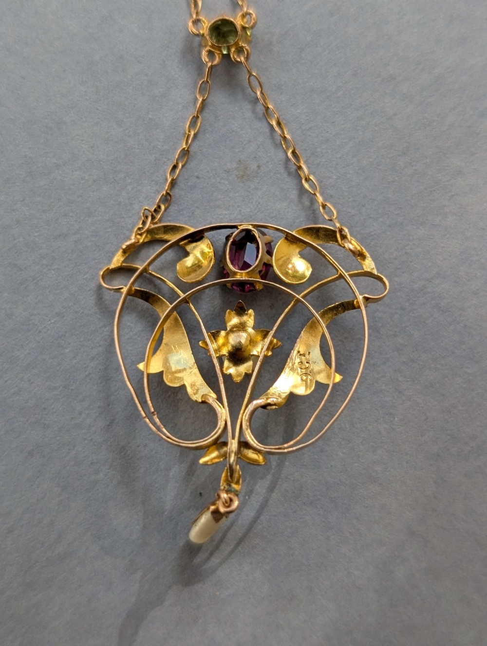 An Edwardian openwork gold lavalier pendant set with an oval purple garnet, a seed pearl center, a pearl drop, and a small green peridot, suspended from a gold chain. View XII