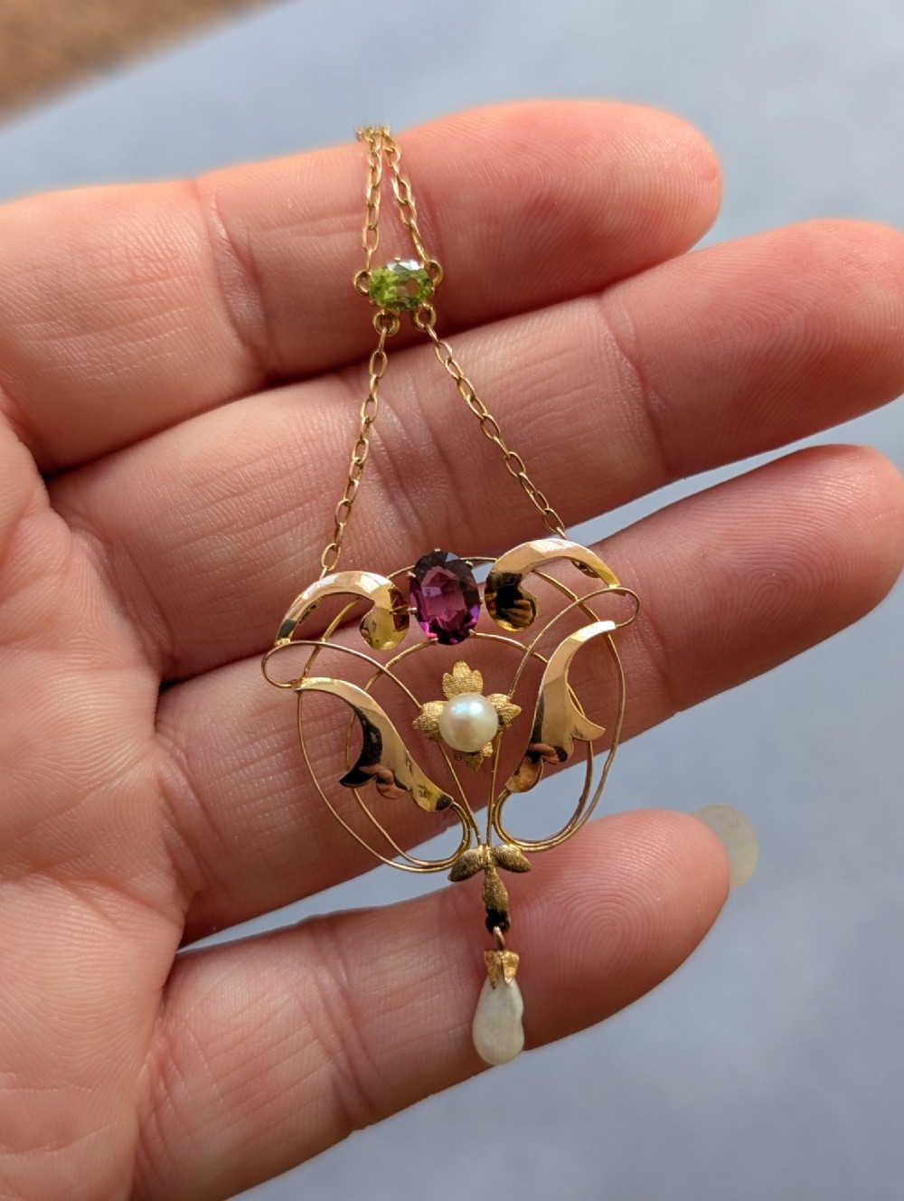 An Edwardian openwork gold lavalier pendant set with an oval purple garnet, a seed pearl center, a pearl drop, and a small green peridot, suspended from a gold chain. View XIII