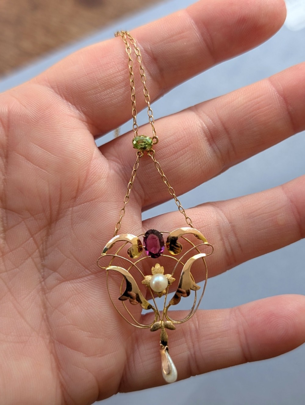 An Edwardian openwork gold lavalier pendant set with an oval purple garnet, a seed pearl center, a pearl drop, and a small green peridot, suspended from a gold chain. View XIV