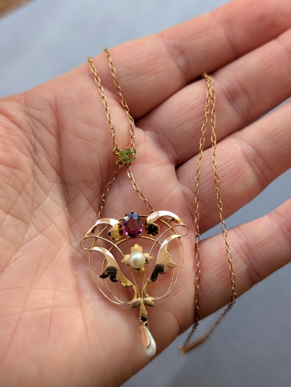 An Edwardian openwork gold lavalier pendant set with an oval purple garnet, a seed pearl center, a pearl drop, and a small green peridot, suspended from a gold chain. View XV