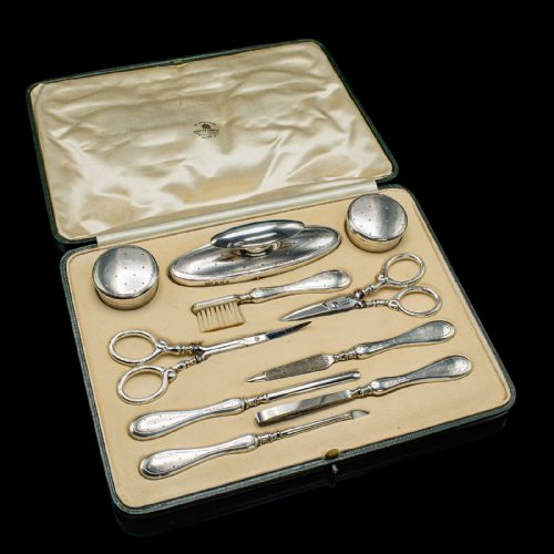 fig 1. An eleven-piece Edwardian silver travelling grooming set arranged against a dark reflective surface, including scissors, nail files, brushes, and small lidded pots with punchwork detailing.