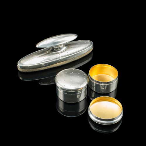 fig 3. An eleven-piece Edwardian silver travelling grooming set arranged against a dark reflective surface, including scissors, nail files, brushes, and small lidded pots with punchwork detailing.