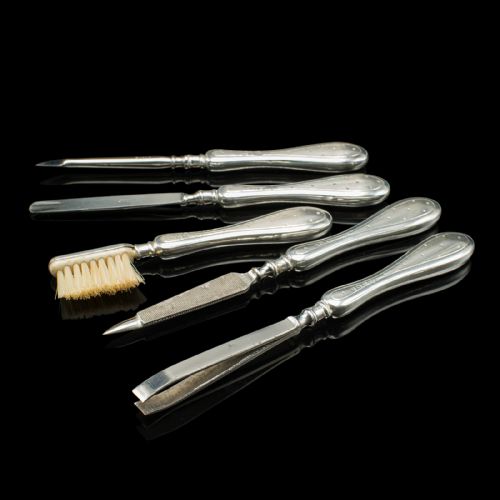 fig 5. An eleven-piece Edwardian silver travelling grooming set arranged against a dark reflective surface, including scissors, nail files, brushes, and small lidded pots with punchwork detailing.