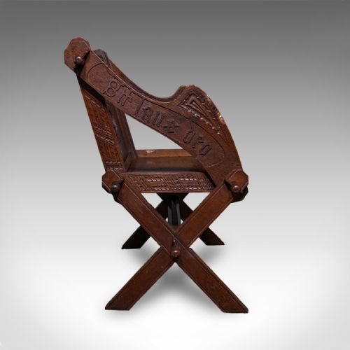 fig 2. A dark oak Glastonbury chair featuring an X-frame structure with wooden pegs, carved Latin inscriptions on the arms, and a carved backrest panel.