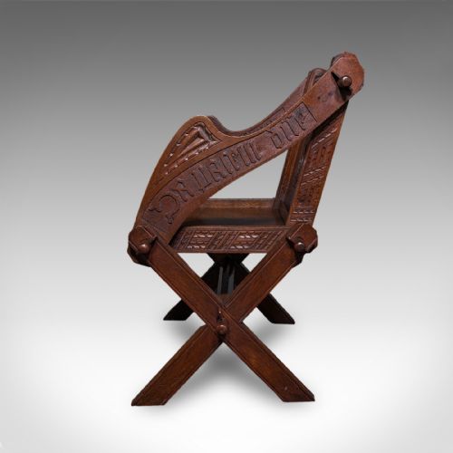 fig 4. A dark oak Glastonbury chair featuring an X-frame structure with wooden pegs, carved Latin inscriptions on the arms, and a carved backrest panel.