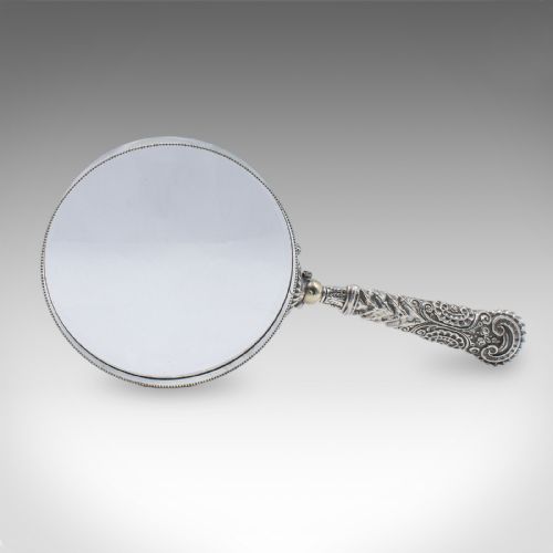 fig 1. An antique Victorian magnifying glass featuring a silver-plated handle with scrollwork relief and a circular optical glass lens with a beaded edge.