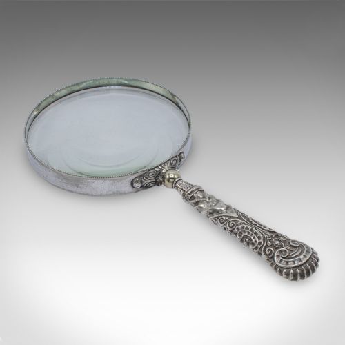 fig 2. An antique Victorian magnifying glass featuring a silver-plated handle with scrollwork relief and a circular optical glass lens with a beaded edge.
