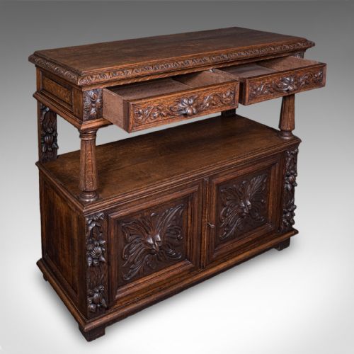 fig 1. A dark Victorian carved oak buffet sideboard featuring a two-tier structure, turned support pillars, carved frieze drawers, and lower cupboard doors decorated with relief carvings of grotesque mask motifs.