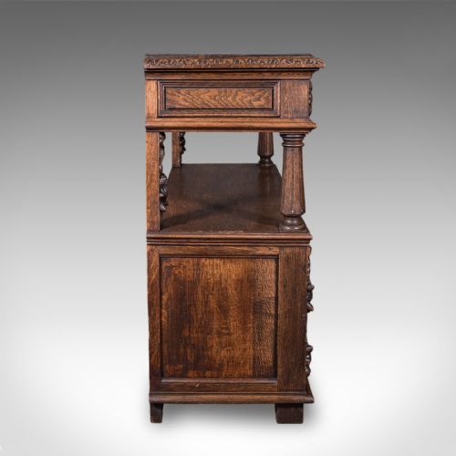 fig 4. A dark Victorian carved oak buffet sideboard featuring a two-tier structure, turned support pillars, carved frieze drawers, and lower cupboard doors decorated with relief carvings of grotesque mask motifs.
