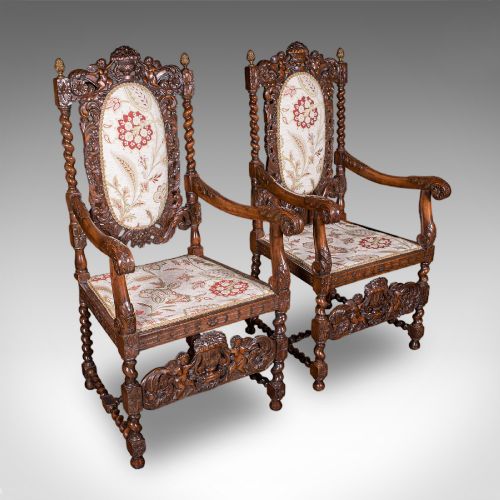 fig 1. A pair of carved dark oak armchairs featuring barley twist uprights, intricate cherub carvings, and cream floral tapestry upholstery with red fringe trim.