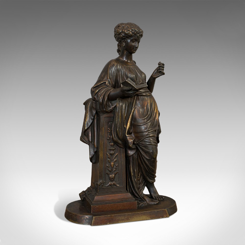 An antique dark bronze figure depicting a draped female maiden holding a book and leaning against a carved pedestal.