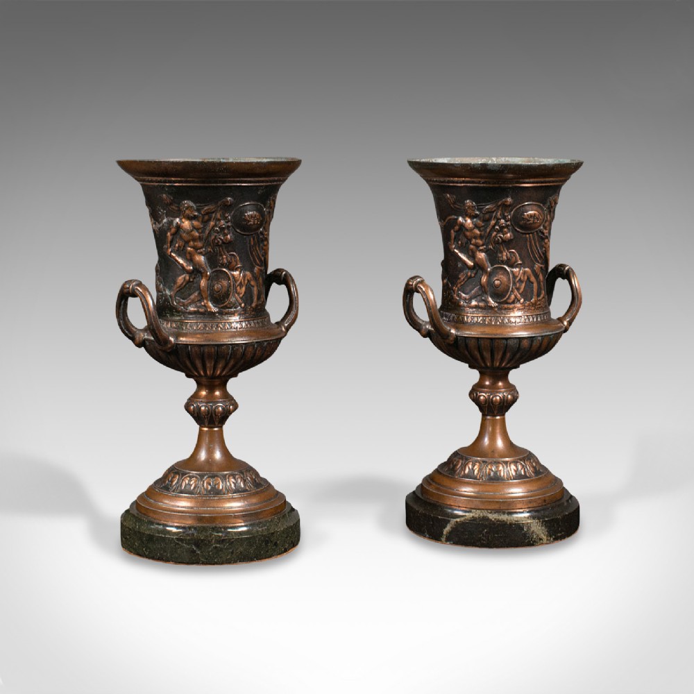 A pair of patinated bronze urns featuring low-relief classical figures set upon dark circular marble bases.