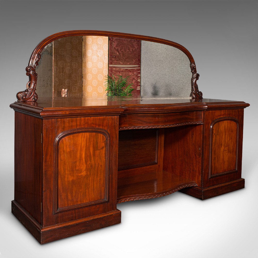 A Victorian flame mahogany pedestal sideboard featuring a large arched mirror back with carved scroll supports above twin panelled pedestal cupboards.