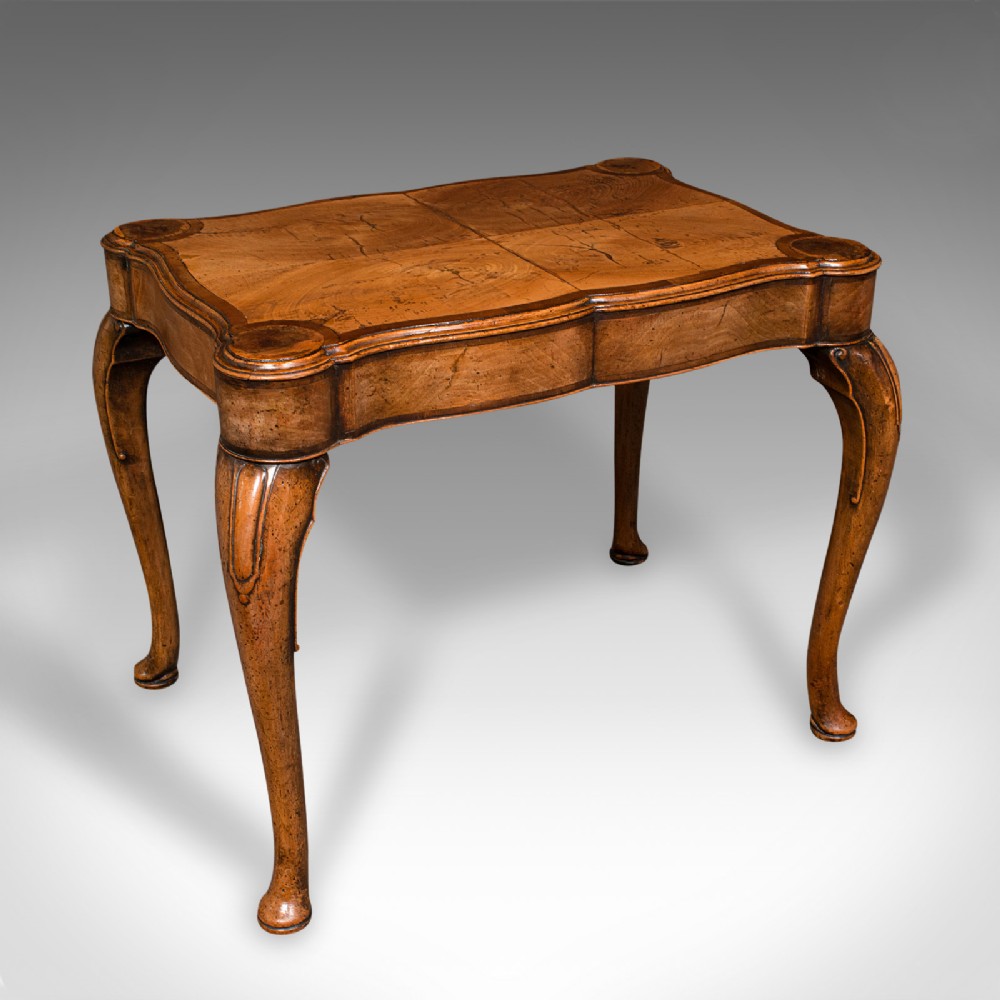 An antique English walnut occasional lamp table featuring a quarter-veneered serpentine top, shaped frieze, and cabriole legs.