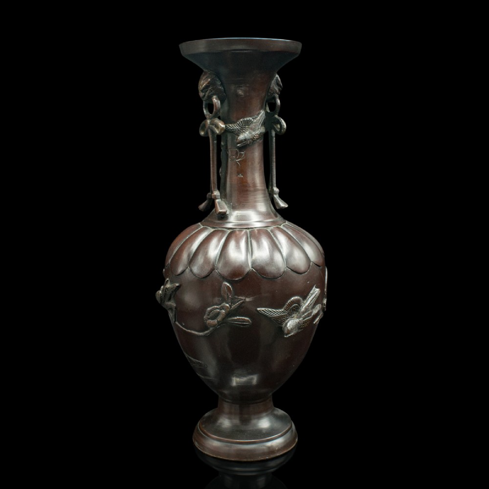 A dark brown patinated bronze stem vase featuring high-relief carvings of birds among foliage, stylised mask handles with drop rings, and a fluted rim.