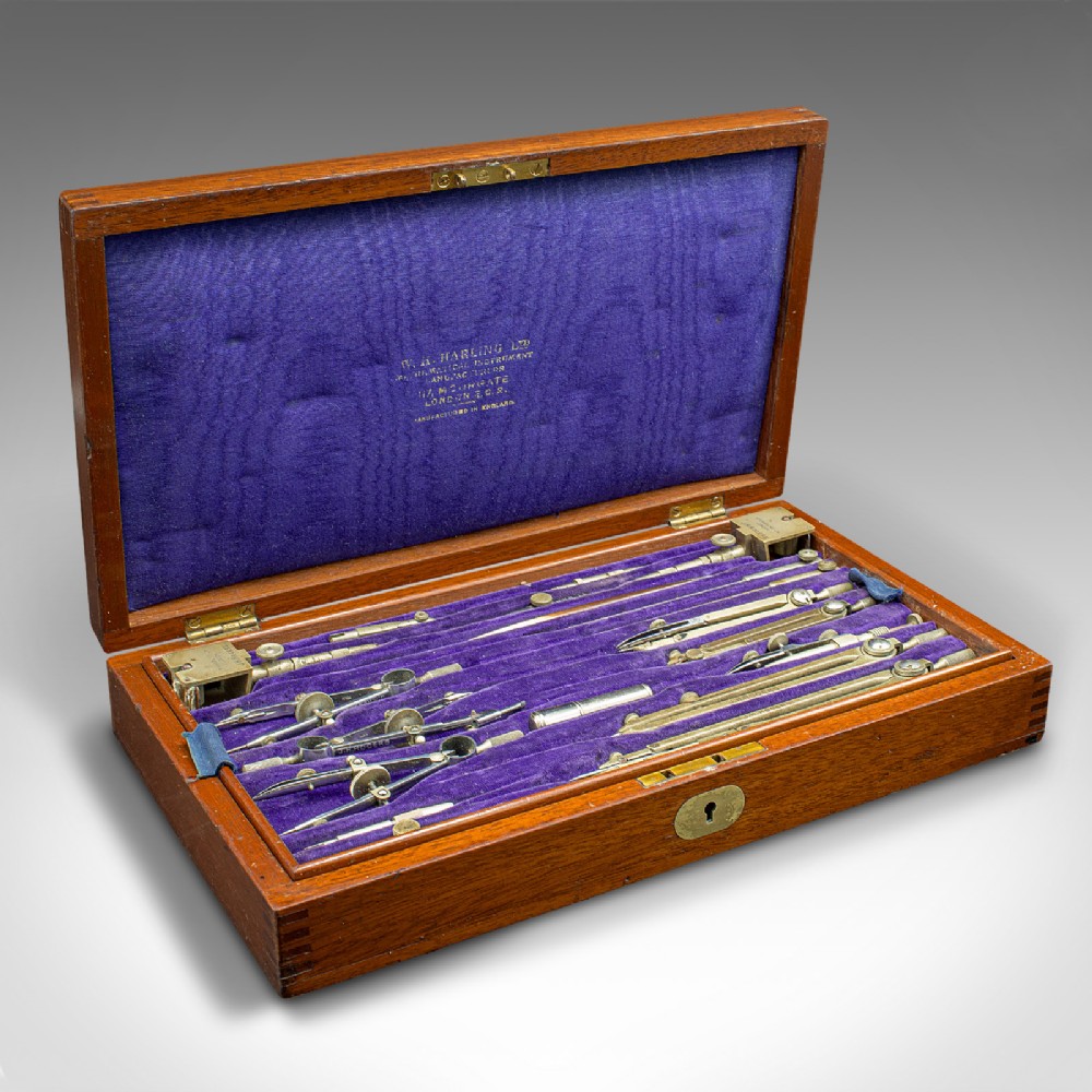 An open mahogany wood box with a purple velvet lining containing an array of antique brass drafting compasses and ruling pens.