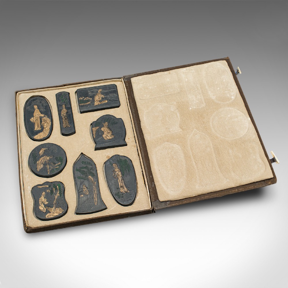 An open brown fabric-lined case containing eight black ink blocks of varied geometric shapes decorated with gilded figurative engravings.